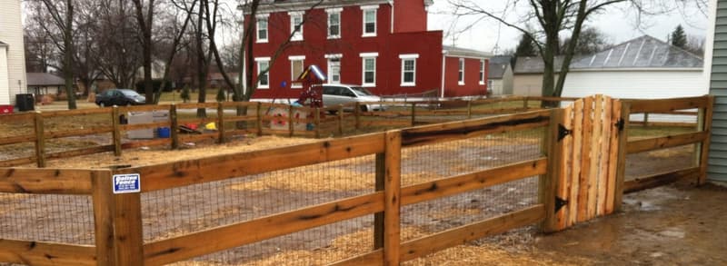 Wood rail fence with wire mesh and gate near red building